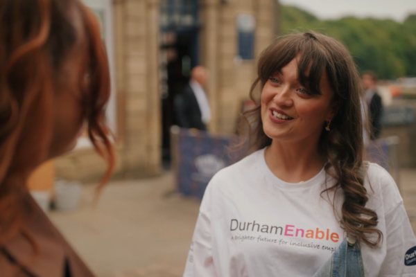 An outdoor scene where a woman in a white “DurhamEnable” T-shirt smiles while talking to someone in the foreground, with a brick building and people in the background.