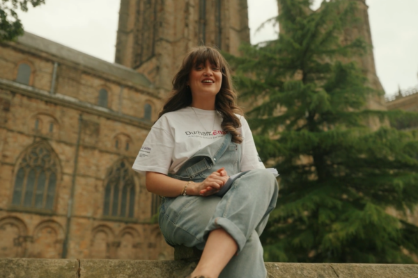 A woman sitting on a stone ledge and smiling in front of Durham Cathedral and a cloudy sky in the background.