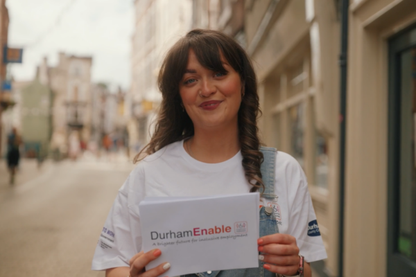 Woman in a white DurhamEnable T-shirt standing on a Durham street, holding a white card with the DurhamEnable logo and text, with buildings and storefronts blurred behind her.